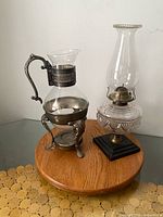 Carafe on metal stand beside oil lamp on round wooden surface