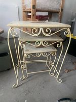 Three cream metal nesting tables stacked, showing scroll legs and floral top