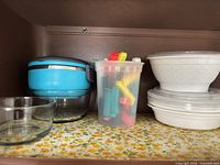 Shelf view showing glass bowl, turquoise chopper bowl, cup of bag clips, stack of white bowls with lids