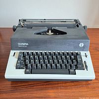 Front view of Olympia Report de Luxe typewriter showing keyboard, logo, model name, and overall color scheme