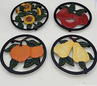 Group view of four round painted cast iron trivets showing sunflower, strawberry, orange and pear motifs