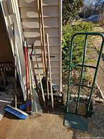 Group of long-handle garden tools and blue push broom leaning against siding next to green hand truck