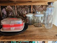 Shelf showing all items: glass canisters with different lids, tall glass bottle, plastic food containers, black round tray, white tray