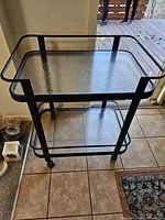 Full view of black metal two-tier bar cart with glass shelves and casters