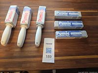 Three handled Pumie toilet bowl ring removers and three Pumie scouring sticks displayed on table