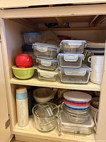 Cupboard shelf with stack of glass containers with blue lids, plastic bowls, measuring cup, additional items