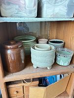 Shelf containing brown glazed planter, sage green rounded planter, tripod beaded planter with beige insert, light gray geometric planter, and sage green classical motif planter