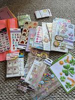 Assorted packs of stickers and embellishments spread on carpet with orange mesh pouch visible