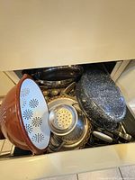 Assorted bakeware and cookware stacked in drawer; visible enamel roaster, red colander, stainless items
