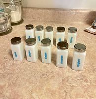Front view of nine milk glass spice jars with blue lettering and metal lids