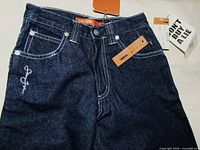 Front view showing dark wash, white stitching, logo embroidery, attached tags