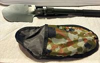 Shovel unfolded beside camouflage pouch showing serrated shovel edge and handle hinge