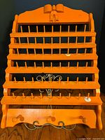 Front angled view of orange wood thread rack showing 57 pegs, metal scroll accent, bow crest