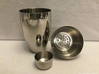 Three-piece stainless steel cocktail shaker disassembled showing body, strainer lid with holes, and measuring cap