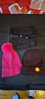 Pair of black gloves, pink pompom beanie, brown beanie displayed on black surface