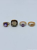 Four rings displayed: purple stone ring, smoky brown cushion ring, purple oval stone ring, rhinestone diagonal band