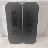 Two black NudeStix metal tins standing upright