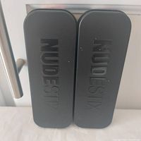 Two closed black metal NudeStix tins, embossed brand name visible