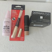 Group shot of all four makeup items – Nudestix stick, NYX spray, Revlon blender in package, Dermablend powder box