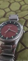 Front angled view of Fossil watch showing red dial, luminous markers, silver hands, stainless steel case and part of bracelet