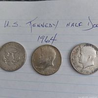 Three 1964 Kennedy half dollars laid out on paper showing both obverse and reverse designs