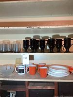 Shelf with smoky grey tumblers, black pedestal mugs, orange mugs, Bloomberg mugs, frosted bowls, white plates