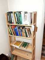 Front view of tall narrow light wood bookcase with two shelves full of books and additional books on lower shelf