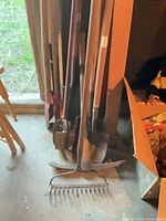 Group of long-handled tools leaning against wall, rake head visible on floor