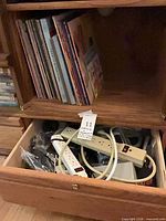 Stack of vinyl LPs in cabinet above open drawer of power strips and cables