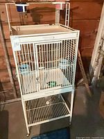 Full view of white metal bird cage on stand with shelf and casters