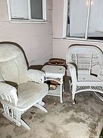 White upholstered glider chair beside wicker table and planter next to wicker rocker