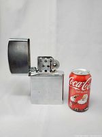 Front view of oversized metal flip-top lighter beside Coca-Cola can for scale