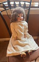 Full view of antique doll seated on chair wearing white dress