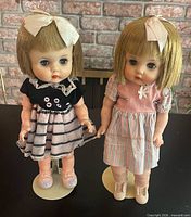 Both Horsman Ruthie dolls standing on cream metal stands, showcasing outfits, shoes and hair ribbons