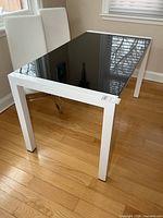 Three-quarter view of table showing white metal frame, black glass top, and extension leaf pull tab