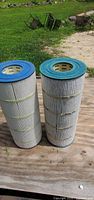 Two Pleatco pool filter cartridges standing upright on wooden surface outdoors