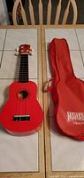 Front view of red Mahalo soprano ukulele beside red gig bag