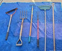 All six garden tools laid out on blanket: squeegee, pitchfork, bow rake, loppers, leaf rake, cultivator