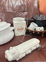Group shot showing Coca-Cola canister, banana hanger, speckled casserole pot, enamel saucepan, mugs, wooden bowl, cow figurines and pig platter