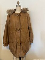 Front view of saddle tan leather coat with fur-trimmed hood showing overall style, pockets and closures