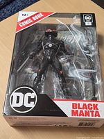 Front view of sealed Black Manta figure in box showing figure, weapons, DC logo