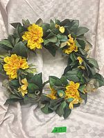 Full view of circular faux wreath with yellow flowers and green leaves against backdrop