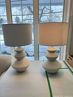 Two matching off-white textured table lamps on table in front of window