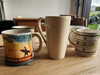 Front view showing three mugs: California Dreaming, tall beige, honeycomb bee pattern with two others partially visible