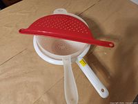 Three kitchen strainers: two handled colanders, one red half-moon strainer