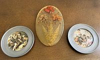 Three decorative pieces: central oval resin plaque with pressed flowers, two round framed porcelain scenic coasters