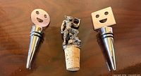 Three bottle stoppers laid out: round smiley face, satyr on cork, square smiley face