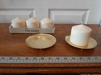 Tray of three packaged pillar candles beside two gold plates and one loose candle, ruler for scale