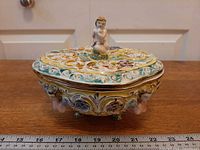 Overall view of footed covered bowl showing cherub finial and side relief detail with ruler for scale