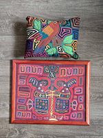 Framed mola artwork and decorative pillow displayed on wooden floor